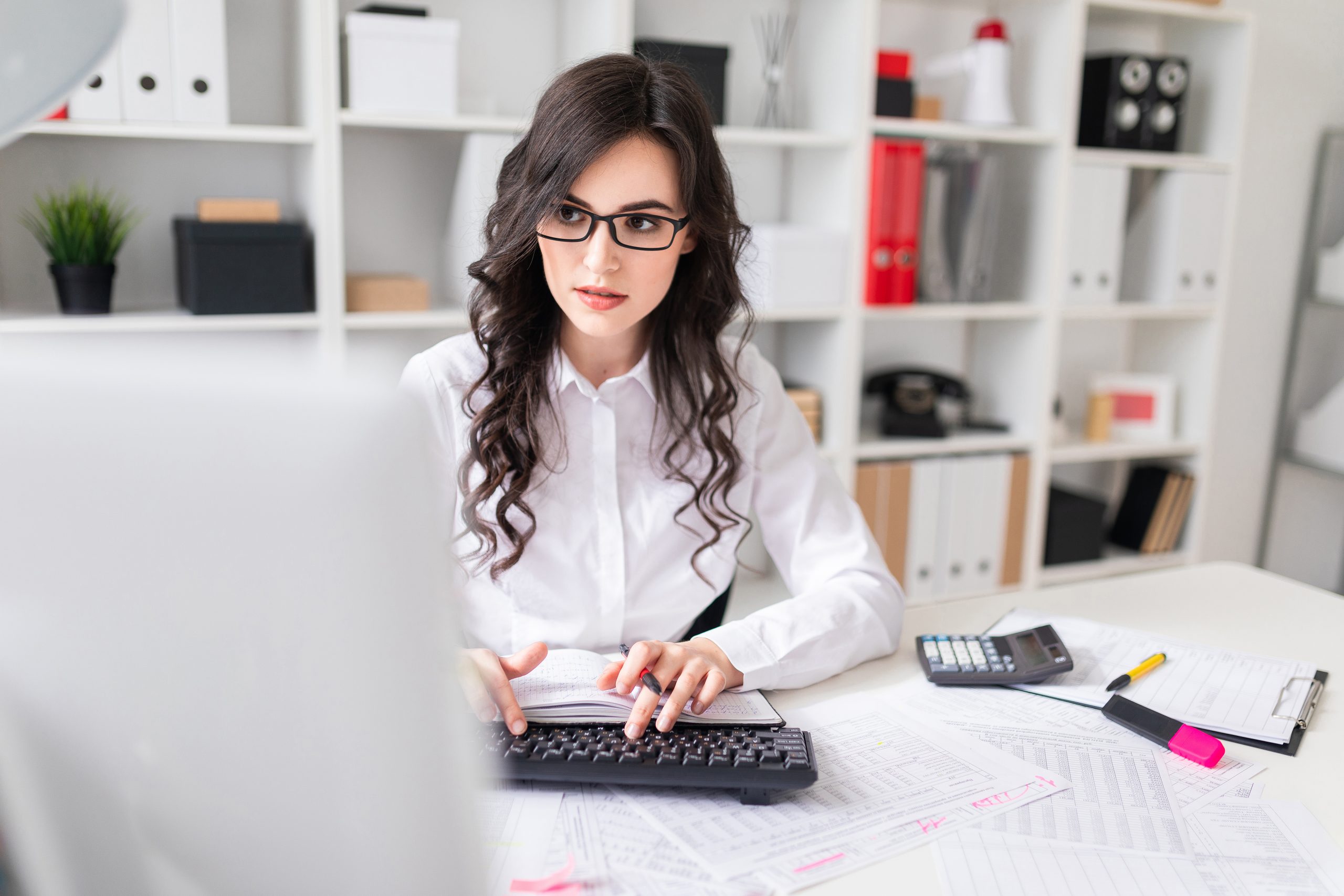 a-young-girl-is-working-at-the-computer-in-the-office-and-holds-a-pen-and-notebook-in-her-hand